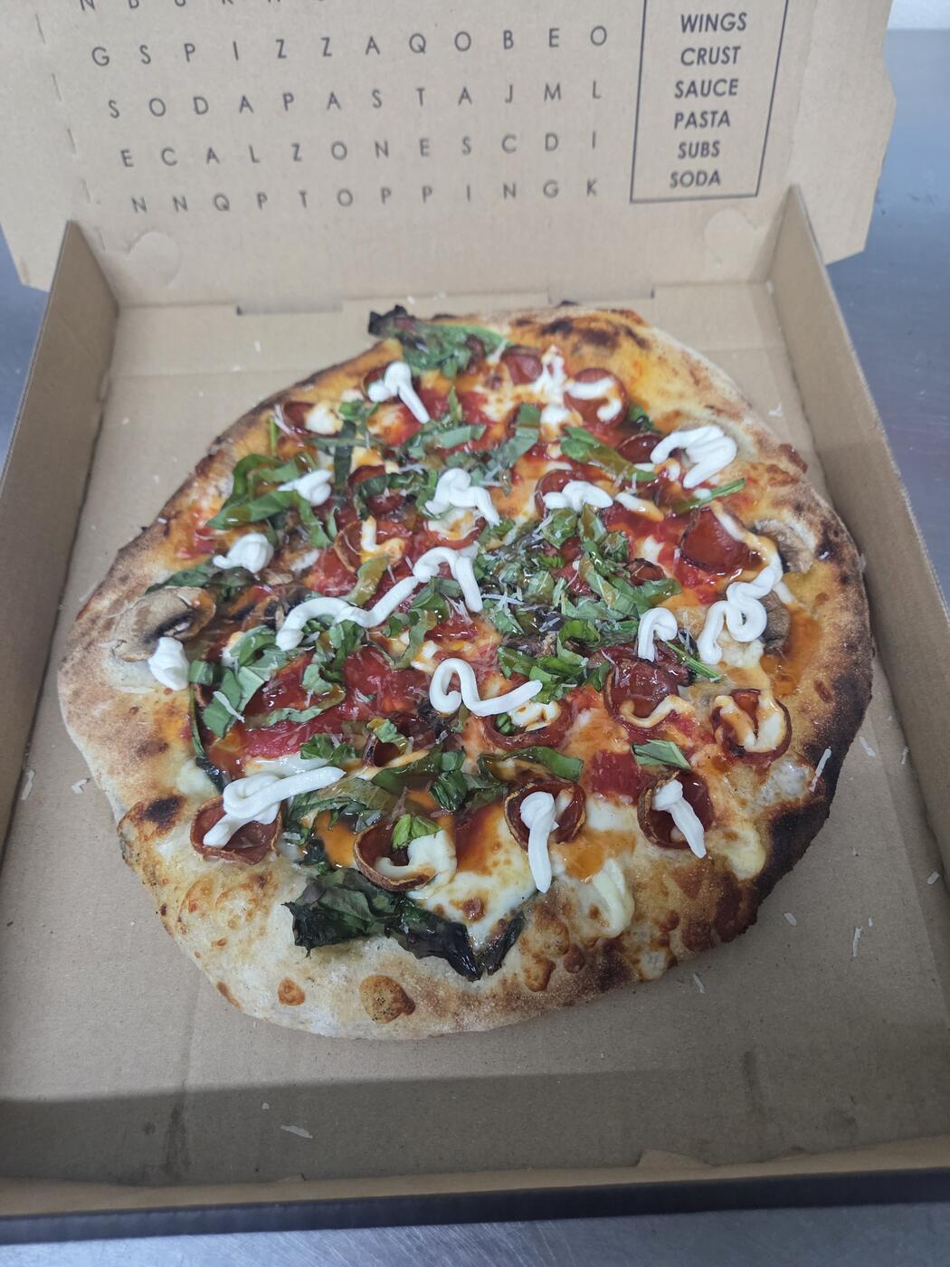 Wood-fired pizza with pepperoni, basil, mushrooms, and a white sauce drizzle